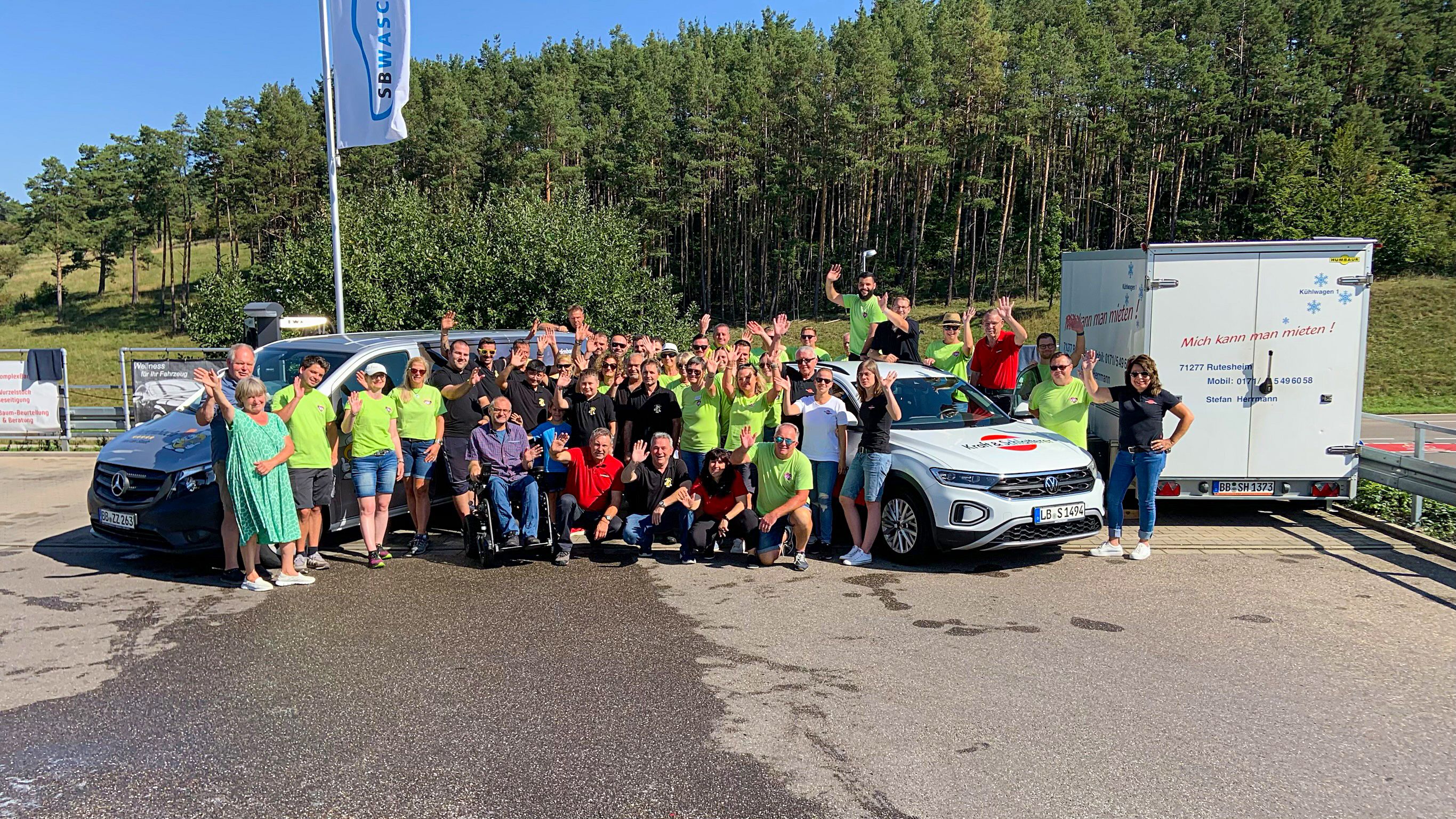Gruppe von Menschen in grünen und schwarzen Shirts posiert fröhlich vor einem weißen Auto und einem weißen Van auf einem gepflasterten Platz, umgeben von Bäumen und Grünflächen.